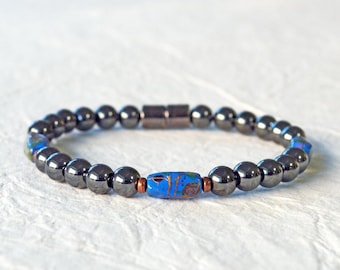 May include: A beaded bracelet featuring dark gray, spherical beads. The bracelet includes a central, rectangular bead with blue, brown, and orange patterns. The clasp is a dark metal. This bracelet is a piece of jewelry.