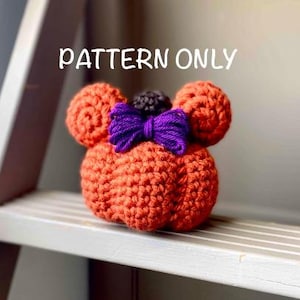 Op de afbeelding: Gehaakte oranje pompoen met twee ronde oren, een paarse strik en een kleine zwarte pompon. De tekst "PATTERN ONLY" staat in het wit boven de pompoen. De pompoen staat op een witte plank.