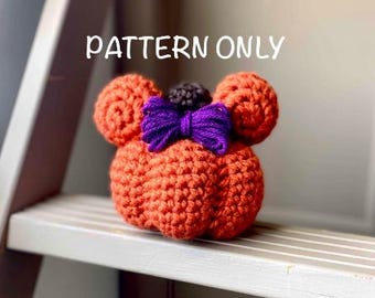 May include: An orange crocheted pumpkin with two round ears, a purple bow, and a small black pom-pom. The text "PATTERN ONLY" is written in white above the pumpkin. The pumpkin is on a white shelf.