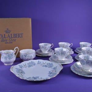 May include: A Royal Albert Bone China tea set featuring a light blue leaf design. The set comprises teacups, saucers, a serving plate, a creamer, and a sugar bowl. The set is presented on a purple backdrop, with the original box visible.