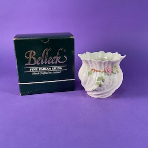 May include: A cream-colored Parian china vase with a scalloped rim and a swirling ribbed texture, featuring a small painted pink rose and green leaves. Beside it sits a dark green box labeled Belleek Fine Parian China Hand Crafted in Ireland.