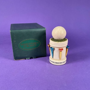 Puede incluir: Un objeto decorativo con temática de golf que presenta una pelota de golf sobre una base cilíndrica con tees de golf coloridos. La base tiene el texto "Golfing Tees of the World". Una caja verde está a la izquierda.