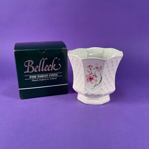 May include: A white Belleek Fine Parian China vase with a basketweave texture and a floral decal featuring pink lilies. The vase is accompanied by its original dark green box, which displays the Belleek logo and the text Fine Parian China.