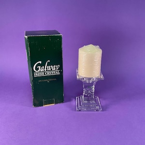 Peut inclure: Un bougeoir en cristal transparent avec une base carrée et une tige torsadée, tenant une bougie crème. La boîte à côté est verte avec le texte "Galway Irish Crystal".