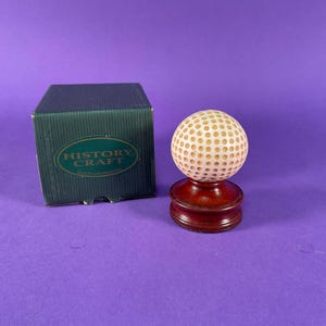 May include: A golf ball replica on a wooden stand, with a dark green box labelled "HISTORY CRAFT" in the background. The golf ball is cream-coloured with small, evenly spaced dimples. The stand is a dark, polished wood.