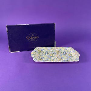 Peut inclure: Un plat de service rectangulaire en céramique avec un motif floral bleu, jaune et blanc, à côté d'une boîte bleu foncé avec "Queen's CHINA". Le plat mesure environ 20 cm de long.