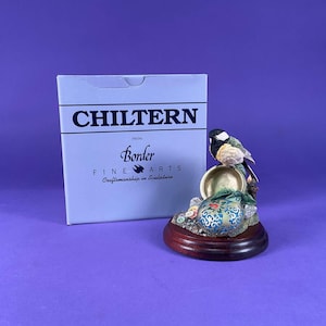 May include: A decorative bird figurine with a black, white, and yellow bird perched on a golden bowl. The figurine sits on a wooden base with a blue and green patterned design. A box with the text "CHILTERN" is in the background.