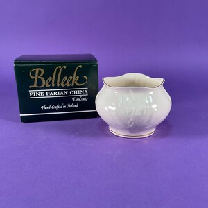 May include: A cream-coloured Belleek Fine Parian China bowl with a scalloped rim and gold trim. The bowl has a floral design and is next to a dark green box with the Belleek logo and text: "Fine Parian China, Hand Crafted in Ireland."