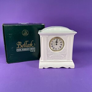 May include: A white ceramic mantel clock with a gold-tone clock face and Roman numerals. The clock is in a rectangular shape with decorative detailing. A green box with the text "Belleek Fine Parian China" is next to the clock.