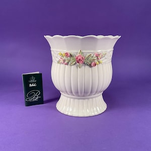 May include: White ceramic planter with a scalloped rim and vertical ribbed design. The planter features a floral garland with pink roses and green leaves. A small card with text is next to the planter.