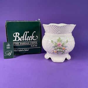 May include: A white Belleek Fine Parian China planter with a floral design, a textured surface, and a scalloped rim. The planter is accompanied by its original box and a small card. The box is green with white text. The planter is hand crafted in Ireland.
