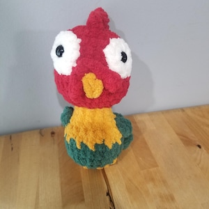 May include: A handmade crochet bird plushie. The bird has a red head with white eyes and black pupils, a yellow beak, and a body of yellow and green. The plushie is sitting on a wooden surface.