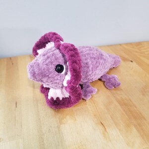 May include: A plush, lavender-colored triceratops dinosaur toy with a dark purple frill and accents. The toy has black button eyes and small, purple feet. The soft, handmade dinosaur is lying on a wooden surface.