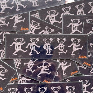 May include: A collection of rectangular stickers featuring dancing bear skeletons in white on a dark background. The stickers have a distressed, vintage look. The artist's signature, "J.Rows," is visible on some stickers.