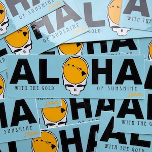May include: A collection of light blue stickers with the word "ALOHA" in large black letters. The "O" is a yellow circle with a map of Hawaii inside a white skull. The text "WITH THE GOLD" and "OF SUNSHINE" are also present.