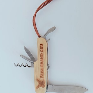 Può includere: Un multiutensile con manico in legno e cinturino in pelle. L'utensile include un coltello, cavatappi, apribottiglie e altri strumenti. Il testo "PAPA GRAND CRU" è visibile sul manico.