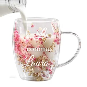 Peut inclure: Une tasse en verre transparente avec une anse, remplie d'un liquide blanc et décorée de fleurs roses et blanches. La tasse porte l'inscription "comme Laura" en écriture cursive. Du lait est versé dans la tasse.