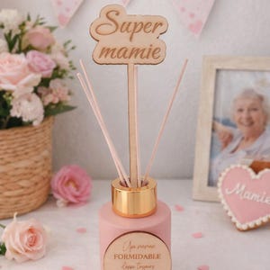 Peut inclure: Un diffuseur rose avec un bouchon doré et des bâtonnets en bois. Un panneau en bois avec l'inscription "Super mamie" est inséré dans le flacon. Le flacon porte une étiquette en bois avec du texte français. Des fleurs roses et une bannière "Bonne fête Mamie" en arrière-plan.