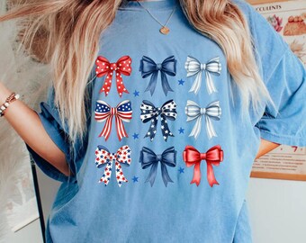 May include: A light blue t-shirt featuring nine bow designs in red, white, and blue. The bows have various patterns, including stars and stripes, and are arranged in three rows of three. The shirt is a casual style.
