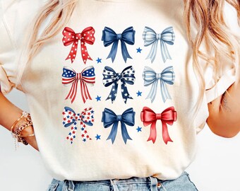 May include: Cream-colored t-shirt featuring a collection of nine decorative bows in red, white, and blue. The bows are arranged in three rows of three, with small blue stars scattered around them. The design evokes a patriotic theme.