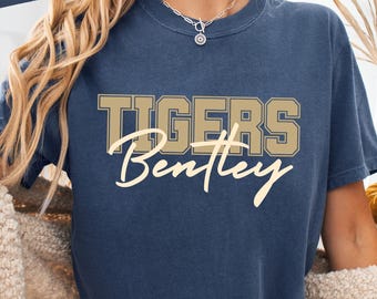 May include: A navy blue t-shirt with the text "TIGERS Bentley" in a gold and cream color scheme. The word "DENIM" is in a navy blue rectangle in the upper left corner. The shirt is worn by a person.