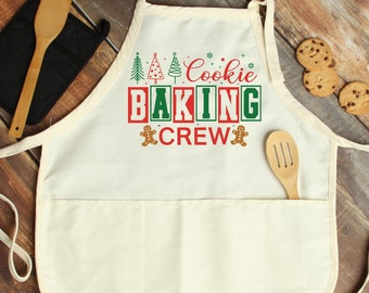 May include: Off-white apron with the text "Cookie Baking Crew" in red and green. The apron has a pocket and is decorated with Christmas tree and gingerbread man graphics. A wooden spatula and oven mitt are visible.