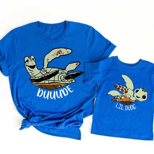 May include: Two blue t-shirts with turtle graphics. The larger shirt has the word "Janna" in white script, and the smaller shirt says "Lil Dude". The turtles are light green, brown, and white.