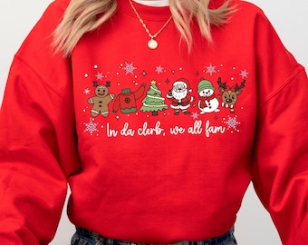 May include: A red sweatshirt featuring a festive design with gingerbread man, sweater, Christmas tree, Santa Claus, snowman, and reindeer characters. The text "In da clerk, we all fam" is printed below the characters.