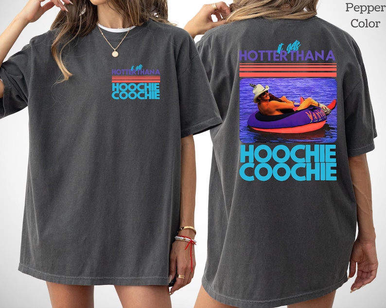 May include: A charcoal gray short-sleeve t-shirt featuring a graphic print. The front displays the text "IT GETS HOTTER THAN A HOOCHIE COOCHIE" in blue and red. The back features the same text with a photo of a person in a hat on a pool float.