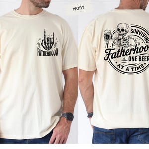 Pu&ograve; includere: T-shirt color avorio con grafiche nere. Il davanti presenta un gesto della mano scheletrica e la parola "FATHERHOOD". Il retro mostra uno scheletro che tiene una birra, con la scritta "SURVIVING FATHERHOOD ONE BEER AT A TIME."