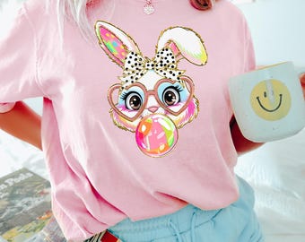 Peut inclure: T-shirt rose avec un lapin de dessin animé portant des lunettes en forme de cœur et soufflant une bulle. Le lapin a un nœud rose et blanc à pois.
