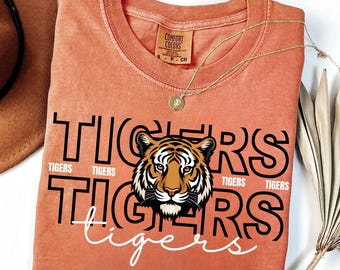 May include: A coral-colored t-shirt with the word "TIGERS" in large black letters and a tiger graphic. The word "TIGERS" is repeated in smaller text. A gold necklace with a pendant is draped on the shirt.