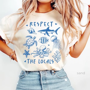May include: A white t-shirt with a blue graphic design that says "Respect the Locals" and features various sea creatures, including a shark, turtle, octopus, starfish, and a jellyfish.