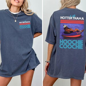 May include: A dark blue t-shirt featuring a graphic on the front left chest and a larger version on the back. The design includes red and teal stripes with the text "HOTTER THAN A HOOCHIE COOCHIE" and an illustration of a person in a tube.
