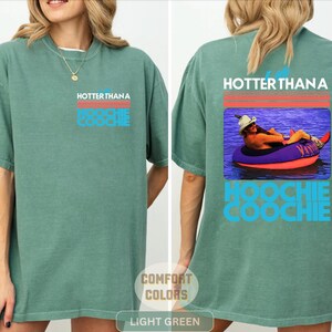 May include: A light green t-shirt featuring a graphic on the front left chest and a larger version on the back. The graphic includes red horizontal lines, the text "HOTTER THAN A HOOCHIE COOCHIE," and a photo of a person in a hat on a pool float.