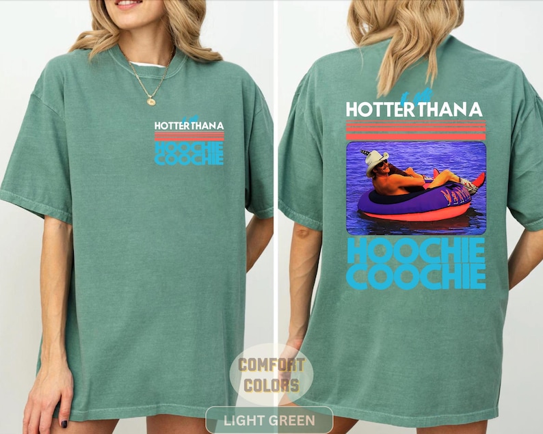 May include: A light green t-shirt featuring a graphic on the front left chest and a larger version on the back. The graphic includes red horizontal lines, the text "HOTTER THAN A HOOCHIE COOCHIE," and a photo of a person in a hat on a pool float.