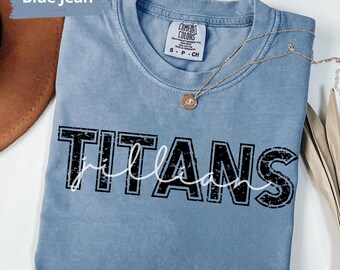 May include: Blue jean colored t-shirt with the word "TITANS" in a distressed black font, with "william" in a white script font underneath. A gold necklace with a pendant is draped across the shirt.