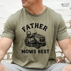May include: Military green t-shirt with the text "FATHER MOWS BEST" above a graphic of a riding lawn mower. The shirt is a solid color and has short sleeves.