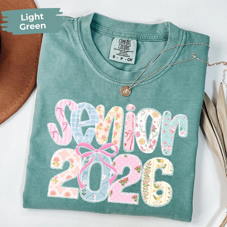 May include: Light green t-shirt with the words "Senior 2026" in a patchwork design. The text is in a mix of floral and patterned fabrics. A silver necklace with a pendant is draped on the shirt.