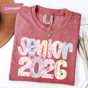 May include: Crimson-coloured t-shirt with the text "Senior 2026" in a patchwork design. The letters and numbers feature floral and patterned fabrics. A silver necklace with a pendant is visible.