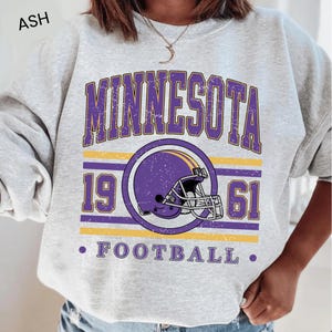 Può includere: Felpa grigio cenere con grafica viola e oro invecchiata. La grafica recita "MINNESOTA" sopra un casco da football viola, con "1961" su entrambi i lati. Sotto c'è la parola "FOOTBALL".
