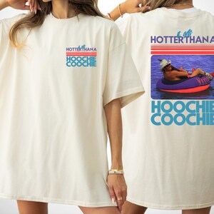 May include: An off-white t-shirt featuring a small graphic on the front and a large graphic on the back. The text reads IT GETS HOTTER THAN A HOOCHIE COOCHIE. The back graphic shows a person in a hat floating on a purple and red inner tube.