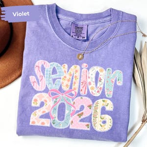May include: A violet Comfort Colors t-shirt featuring the text "Senior 2026" in a patchwork design. The design incorporates floral and striped patterns in pink, blue, and green. A gold necklace with a pendant rests on the shirt.
