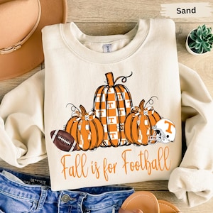 May include: Sand-colored sweatshirt with a fall-themed graphic. The design features three orange pumpkins, a football, and a football helmet. The text "Fall is for Football" is printed in orange.