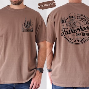 Pu&ograve; includere: T-shirt color espresso con grafica di mano di scheletro e la scritta "Fatherhood" sul davanti. Il retro presenta uno scheletro con una birra e la scritta "Surviving Fatherhood One Beer at a Time."