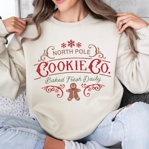 May include: A cream-colored sweatshirt with a festive design. The graphic reads "North Pole Cookie Co. Baked Fresh Daily" in red and green, with snowflake and gingerbread man accents. The sweatshirt is a cozy, casual garment.