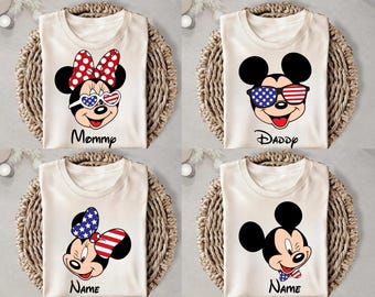 May include: Four cream-colored t-shirts with Mickey and Minnie Mouse designs. Each shirt features a different design, including Mickey and Minnie with patriotic sunglasses and bows. The shirts are arranged on woven wicker placemats.