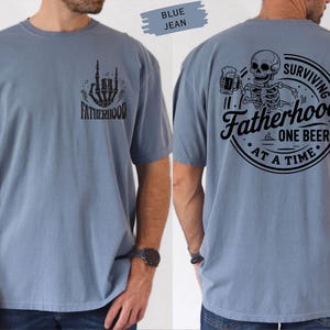 Pu&ograve; includere: T-shirt blu jeans a maniche corte con grafiche nere. Il davanti presenta una mano di scheletro e la scritta "FATHERHOOD". Il retro mostra uno scheletro con una birra e la scritta "SURVIVING FATHERHOOD ONE BEER AT A TIME".