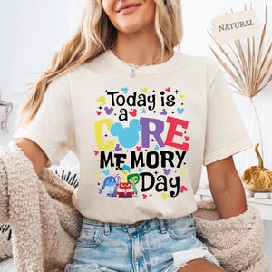 May include: A white t-shirt featuring a colourful graphic that reads "Today is a CORE MEMORY Day" with a Mickey Mouse head in the centre of the word "CORE". The graphic also includes five characters from the film Inside Out.