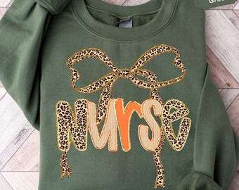May include: Military green sweatshirt with a leopard print bow and the word "NURSE" in a combination of leopard print, orange, and tan. The sweatshirt has long sleeves and a crew neck.